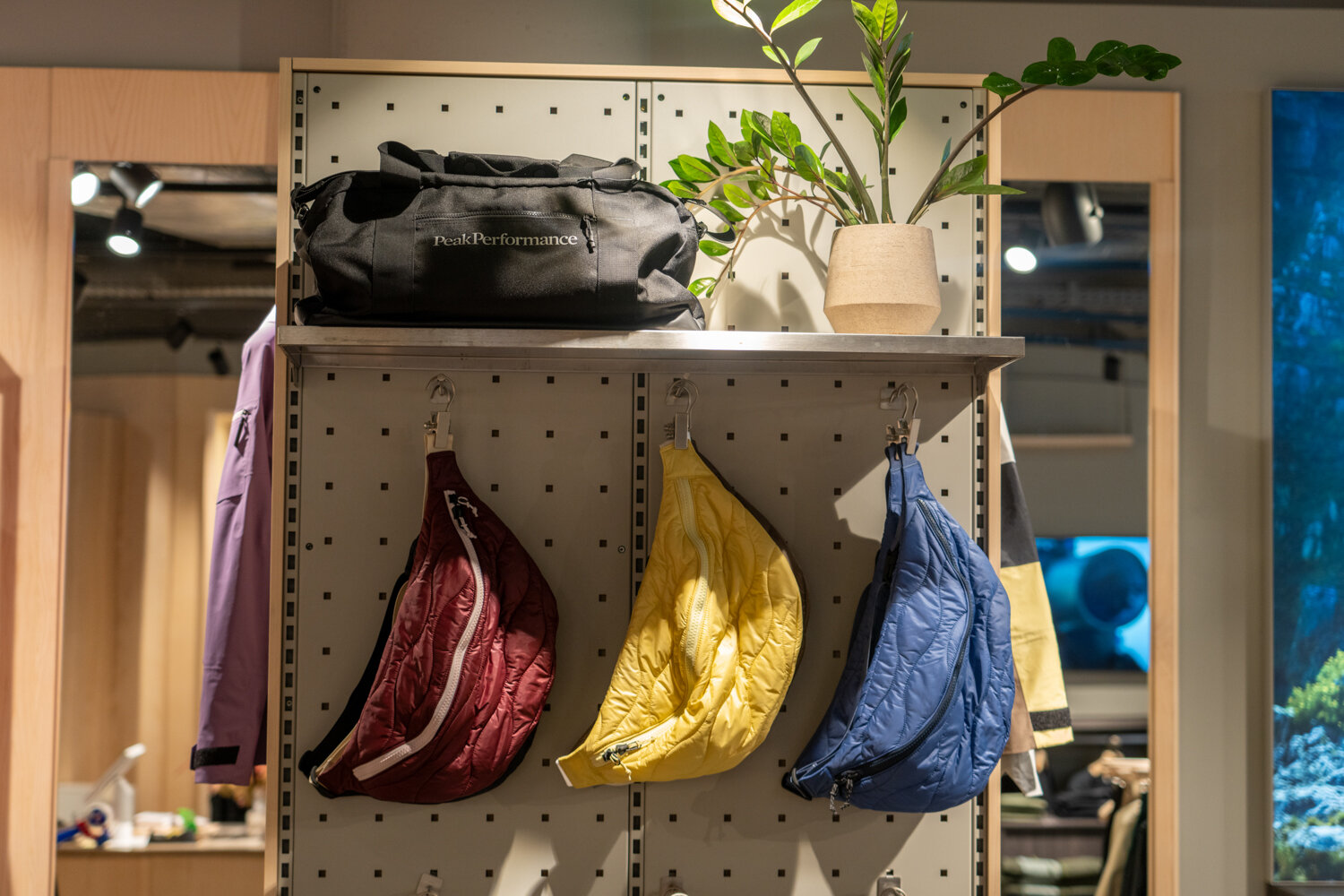 Lochwand bei Peak Performance mit gehängten Bauchtaschen in Weinrot, Gelb und Blau sowie einer schwarzen Reisetasche auf der Ablage.