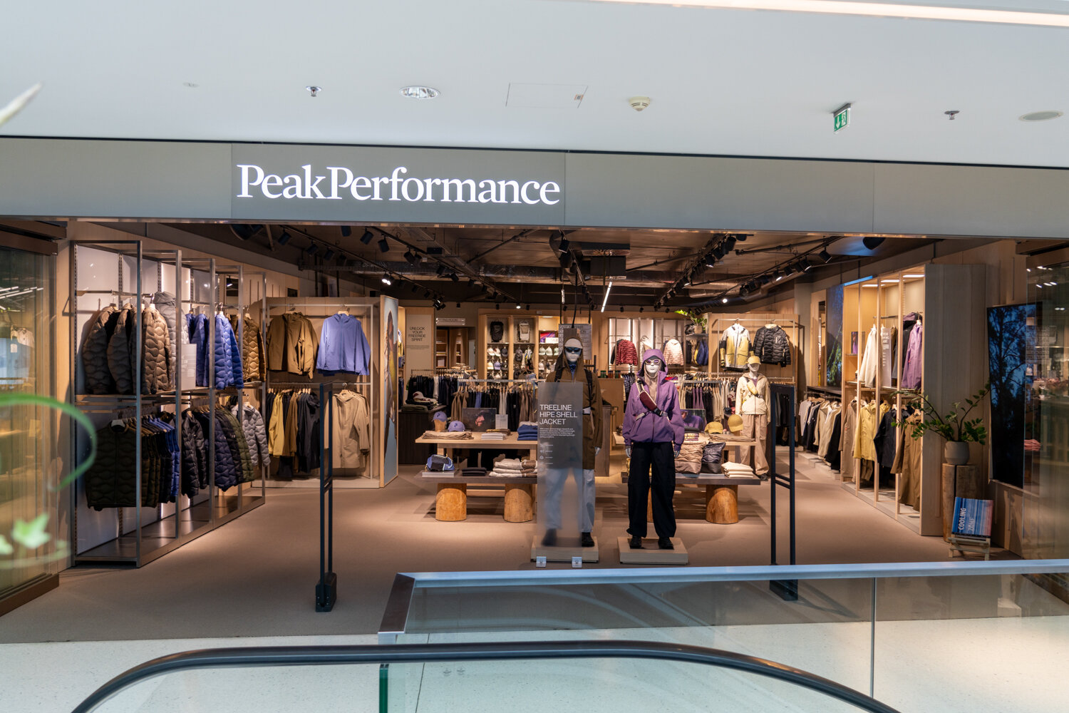 Offene Eingangsfront des Peak Performance Stores im Einkaufszentrum mit beleuchtetem Logo, Schaufensterpuppen und Blick auf Outdoor-Bekleidung.