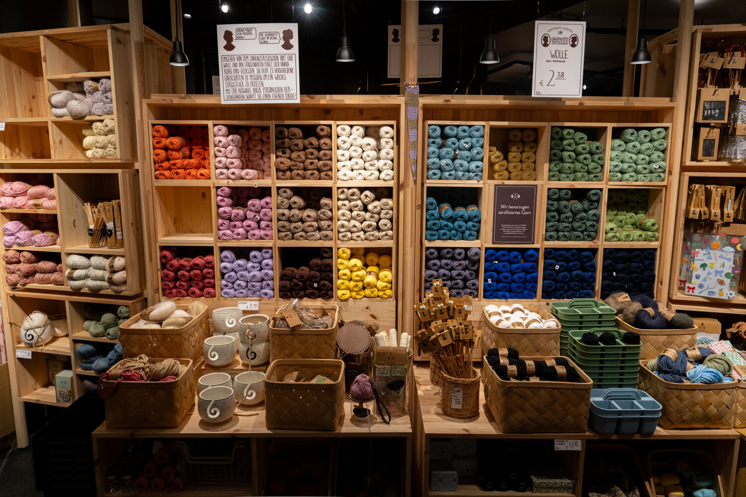 Helle Holzregale bei Søstrene Grene gefüllt mit farblich sortierter Wolle, Garnknäueln und Flechtkörben mit Handarbeitszubehör.