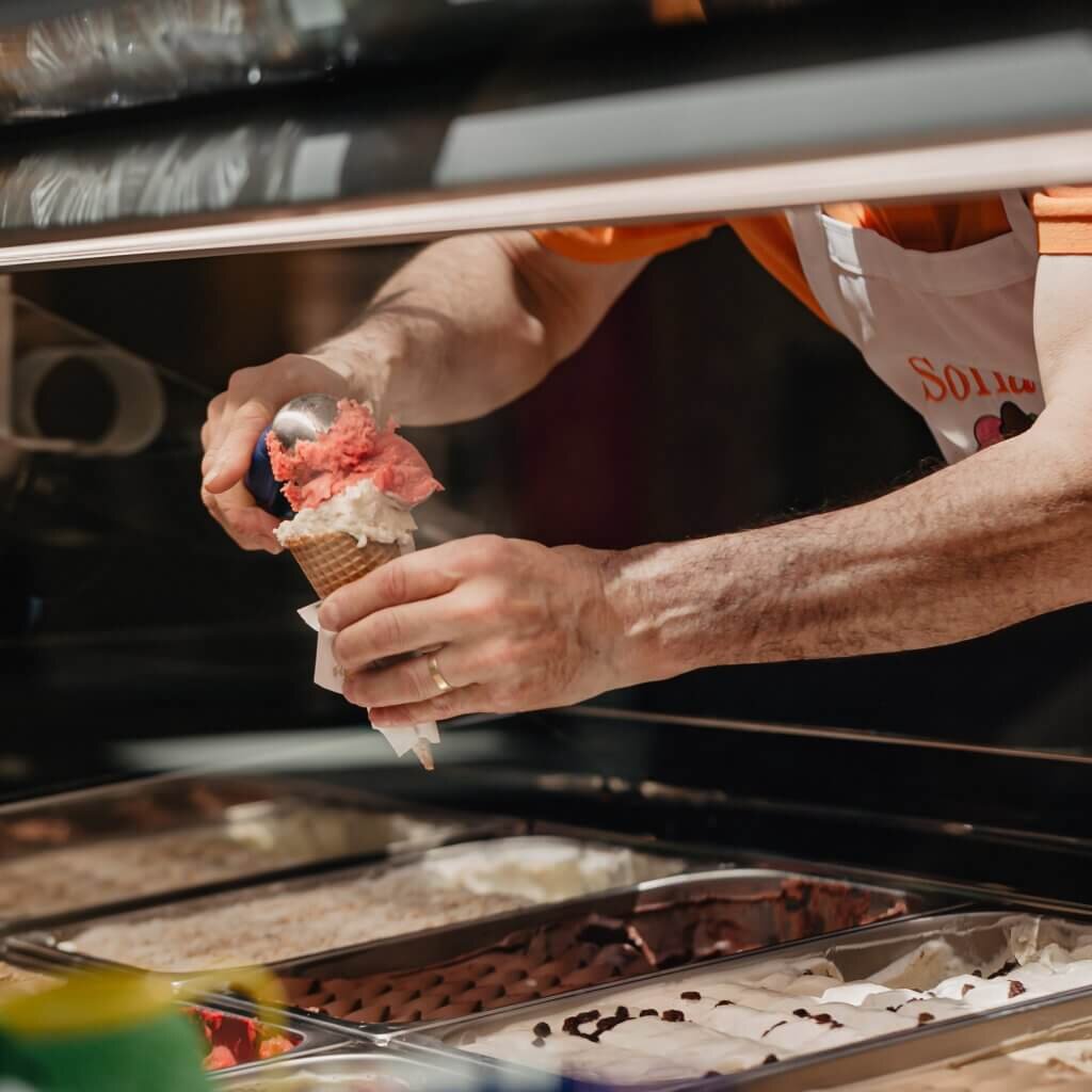 Ein Mitarbeiter portioniert mit einem Eisportionierer frisches, fruchtiges Eis direkt in eine knusprige Waffel über den Edelstahl-Eisbehältern.