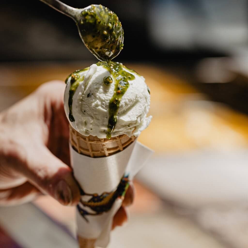 Nahaufnahme einer Eiswaffel mit cremigem weißen Eis, über das mit einem Löffel eine feine, grüne Pistazien-Sauce gegossen wird.