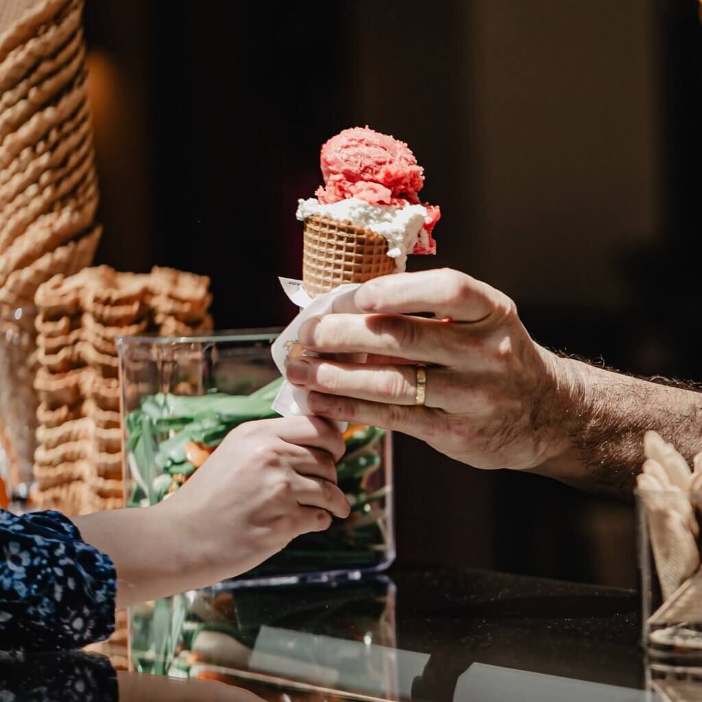 Detailaufnahme der Übergabe einer frisch zubereiteten Eiswaffel mit zwei Kugeln Eis von der Bedienung an einen Kunden.