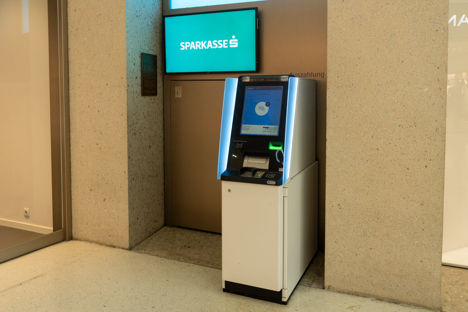 Seitliche Perspektive des Sparkasse-Bankomaten mit leuchtendem Bildschirm und Sparkasse-Logo an der Wand.