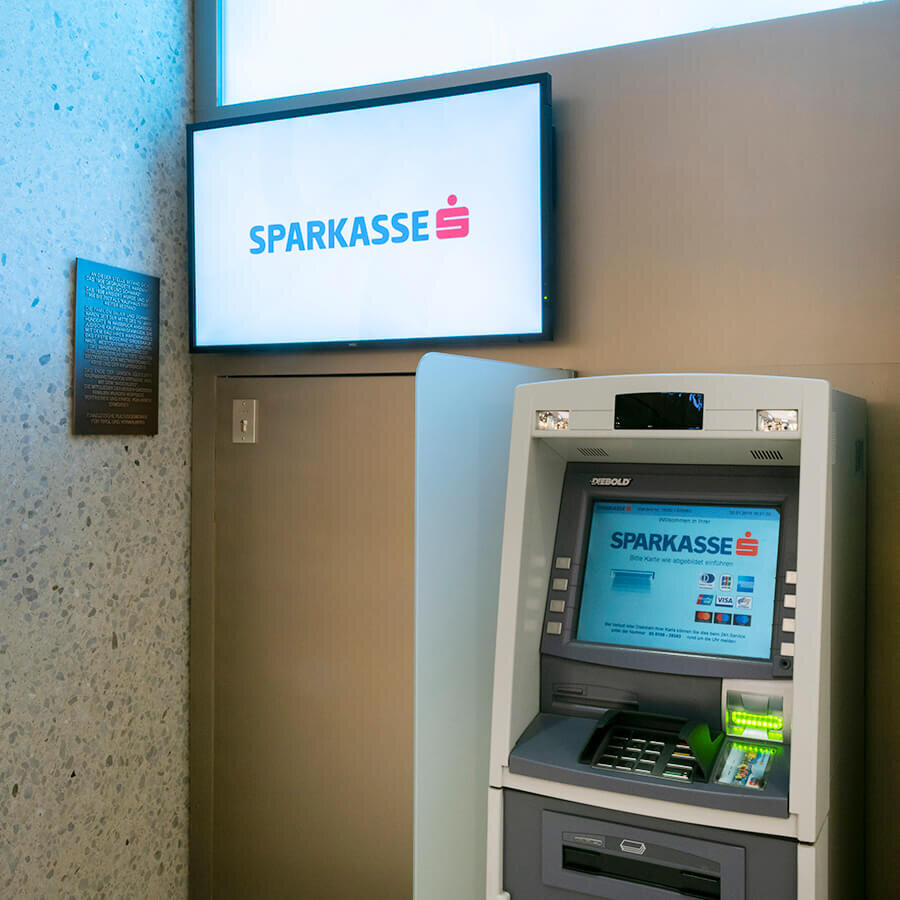 Ein moderner Geldausgabeautomat (Bankomat) der Sparkasse mit einem Infobildschirm und einer historischen Gedenktafel an der Wand.