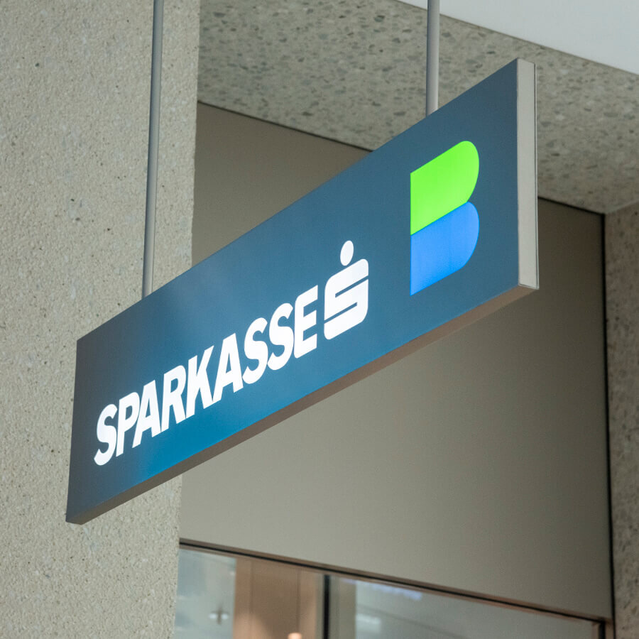 Ein beleuchtetes, von der Decke hängendes blaues Hinweisschild mit dem weißen Schriftzug „SPARKASSE“ und einem grün-blauen Logo-Element.