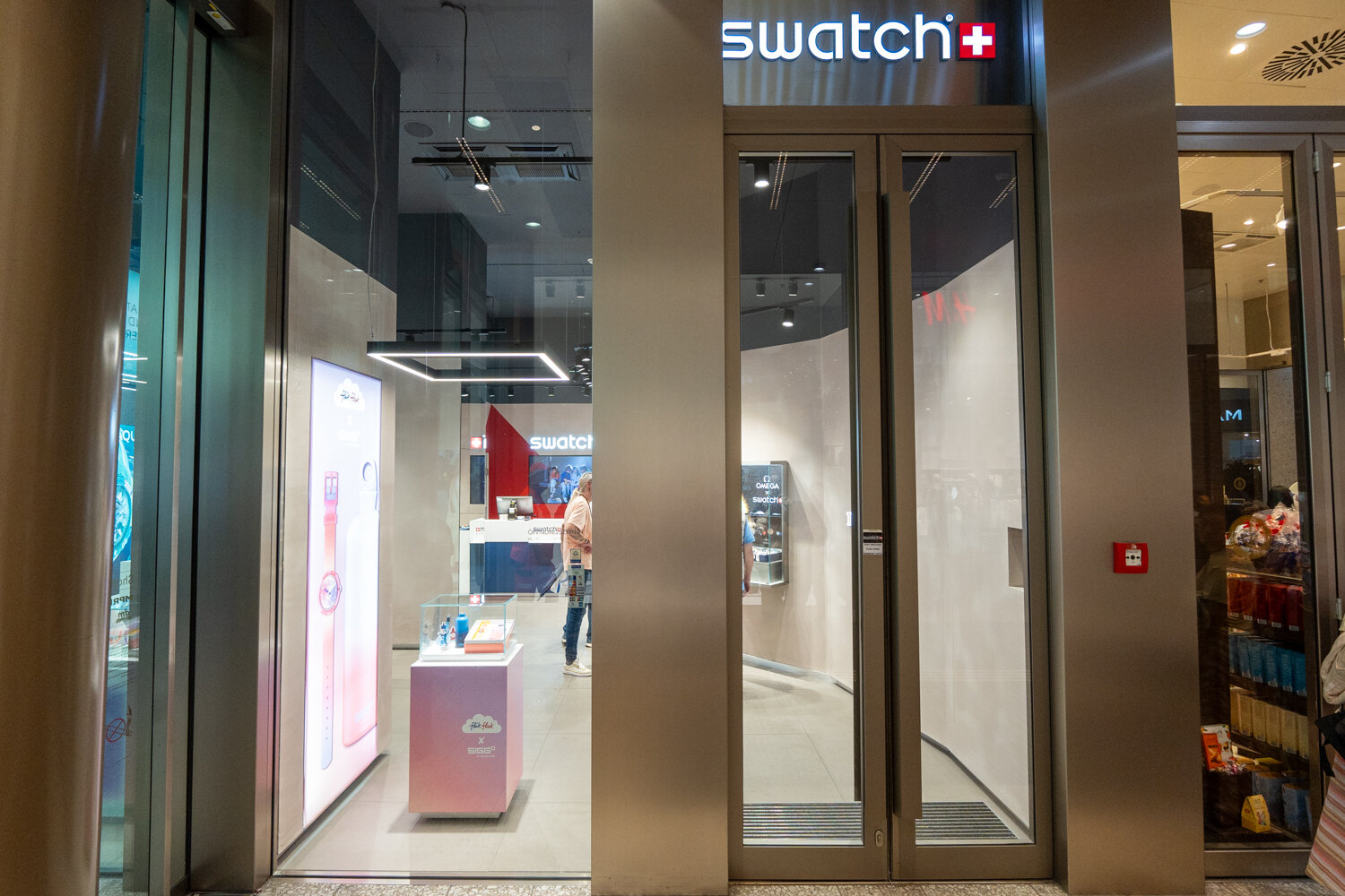 Eingangsbereich und Schaufenster eines Swatch Stores im Einkaufszentrum mit beleuchtetem Markenlogo und Uhren-Displays.
