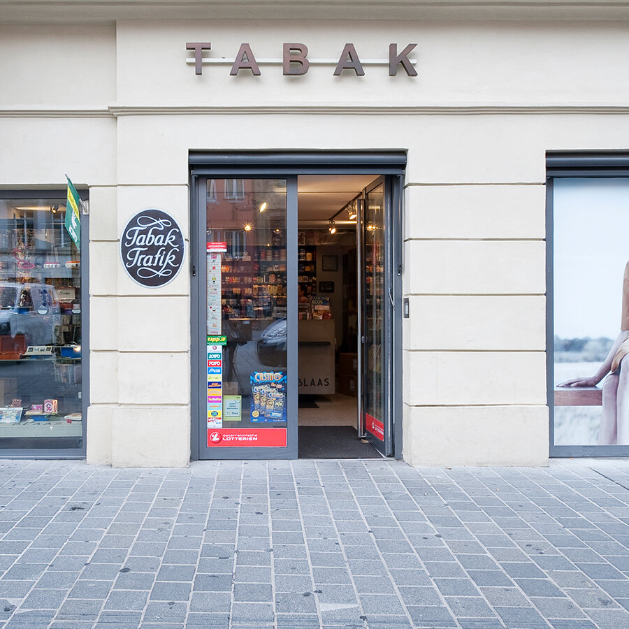 Frontalansicht des Eingangs der Trafik Blaas mit dem metallenen Schriftzug „TABAK“ an der hellen Fassade und Lotterie-Logos an der Glastür.