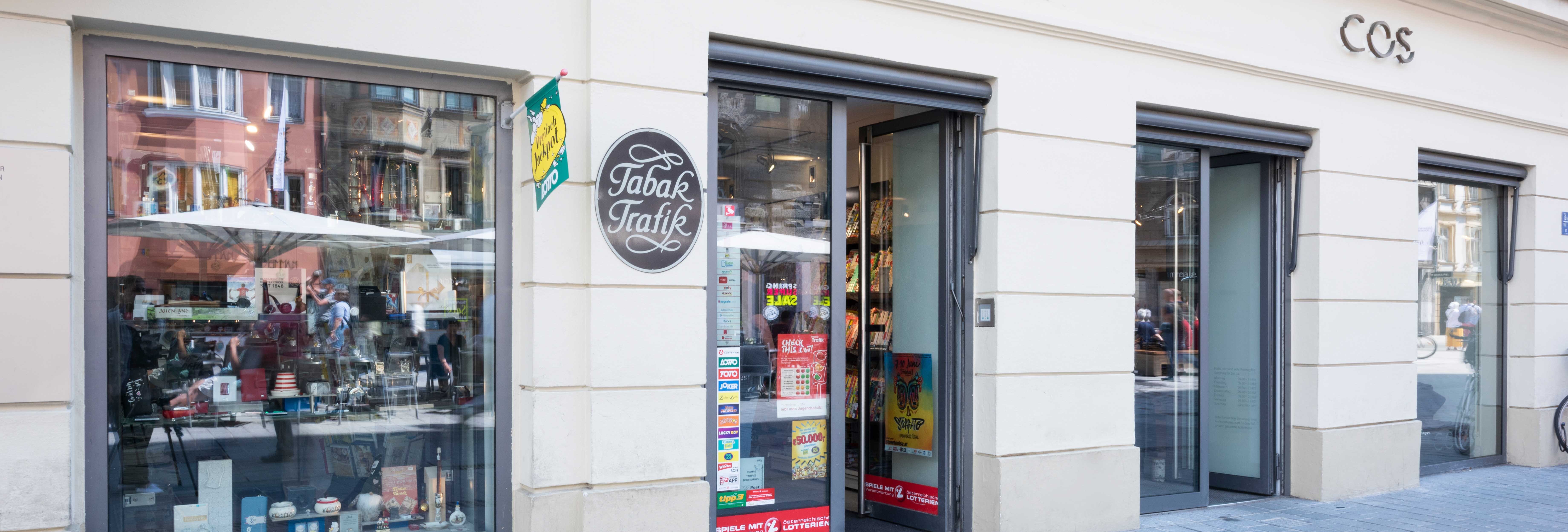Weitläufiges Breitbild-Panorama der Ladenzeile mit dem einsehbaren Schaufenster der Trafik Blaas und dem angrenzenden COS Modegeschäft.