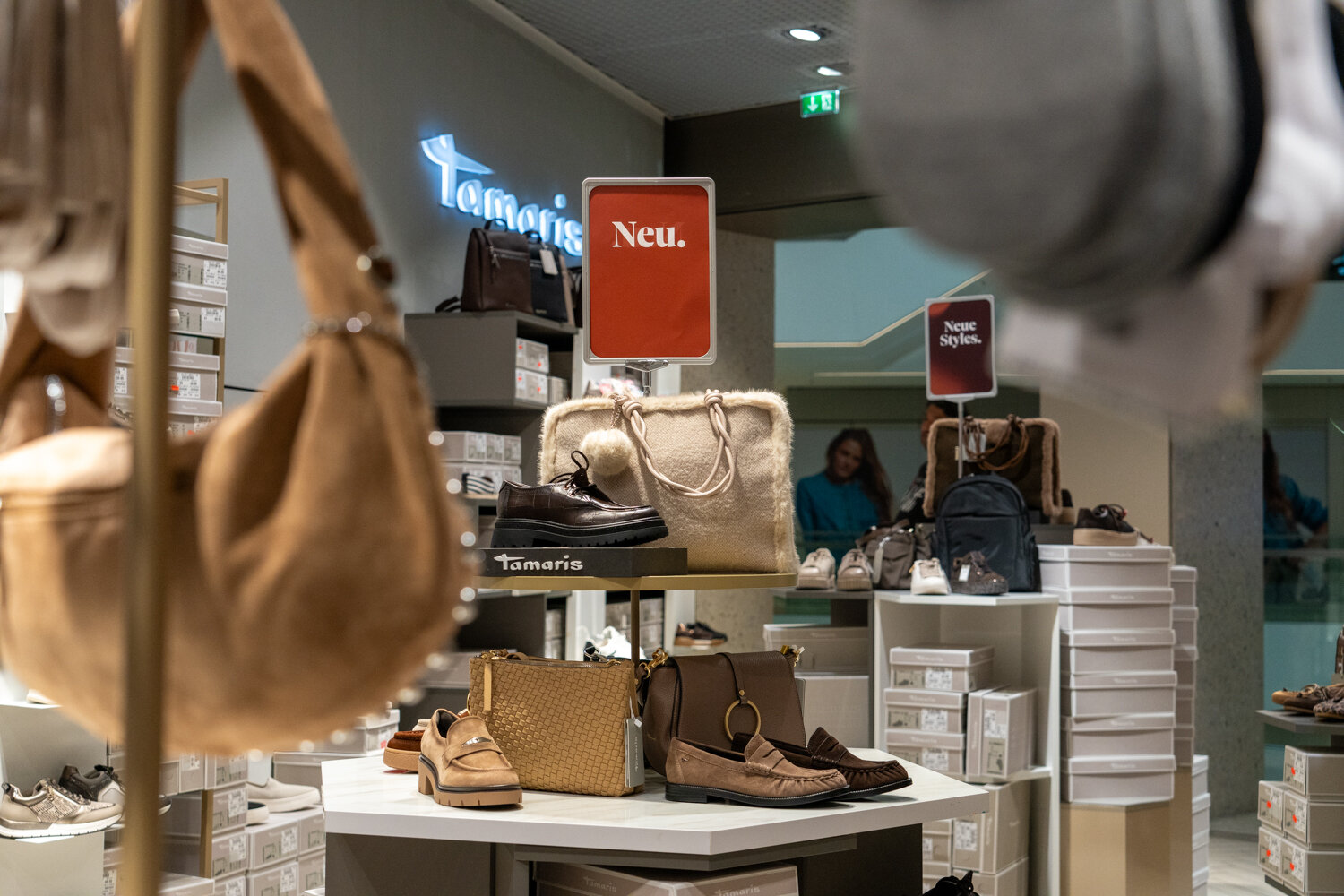 Verkaufsdisplay bei Tamaris mit Damen-Schuhen, Handtaschen und Rucksäcken unter dem Schild Neu.