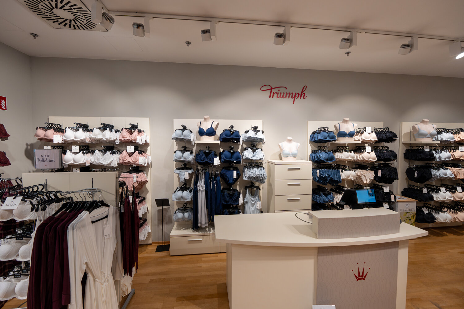 Übersichtsaufnahme des Triumph Geschäfts mit Beratungstresen, Markenlogo an der Wand und Kleiderständern für Lingerie.