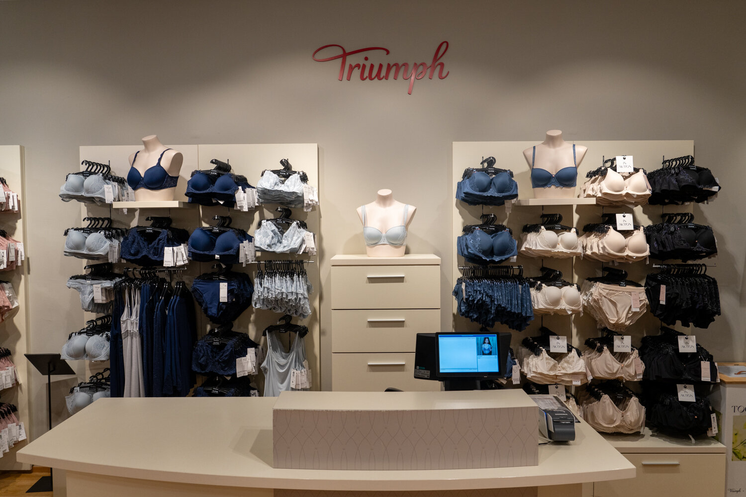 Kassenbereich im Triumph Store mit weißem Tresen, Bildschirm und dahinter stehenden Wandregalen voll Damenunterwäsche.