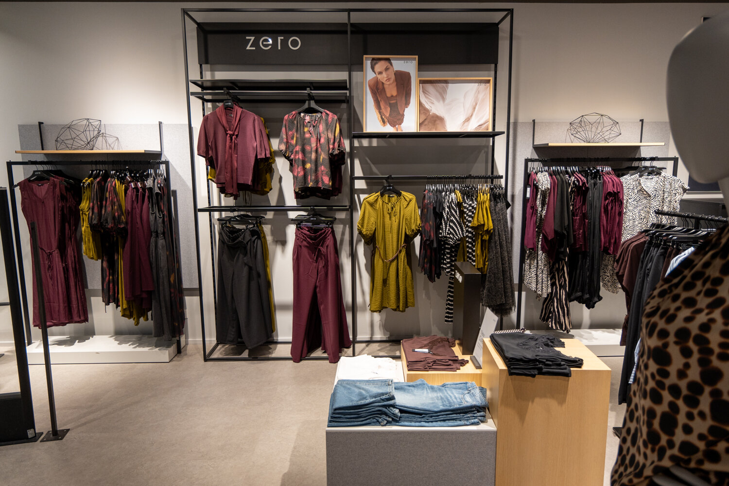 Wanddisplay im zero Store mit beerenfarbenen und senfgelben Outfits, Hosen, Blusen und Stapeln von Jeans.
