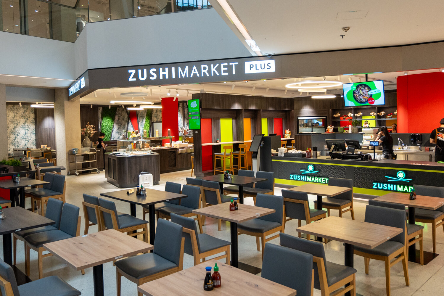 Weitwinkelaufnahme des Zushi Market Plus Restaurants mit zahlreichen Holztischen, Stühlen und Buffetbereich im Hintergrund.
